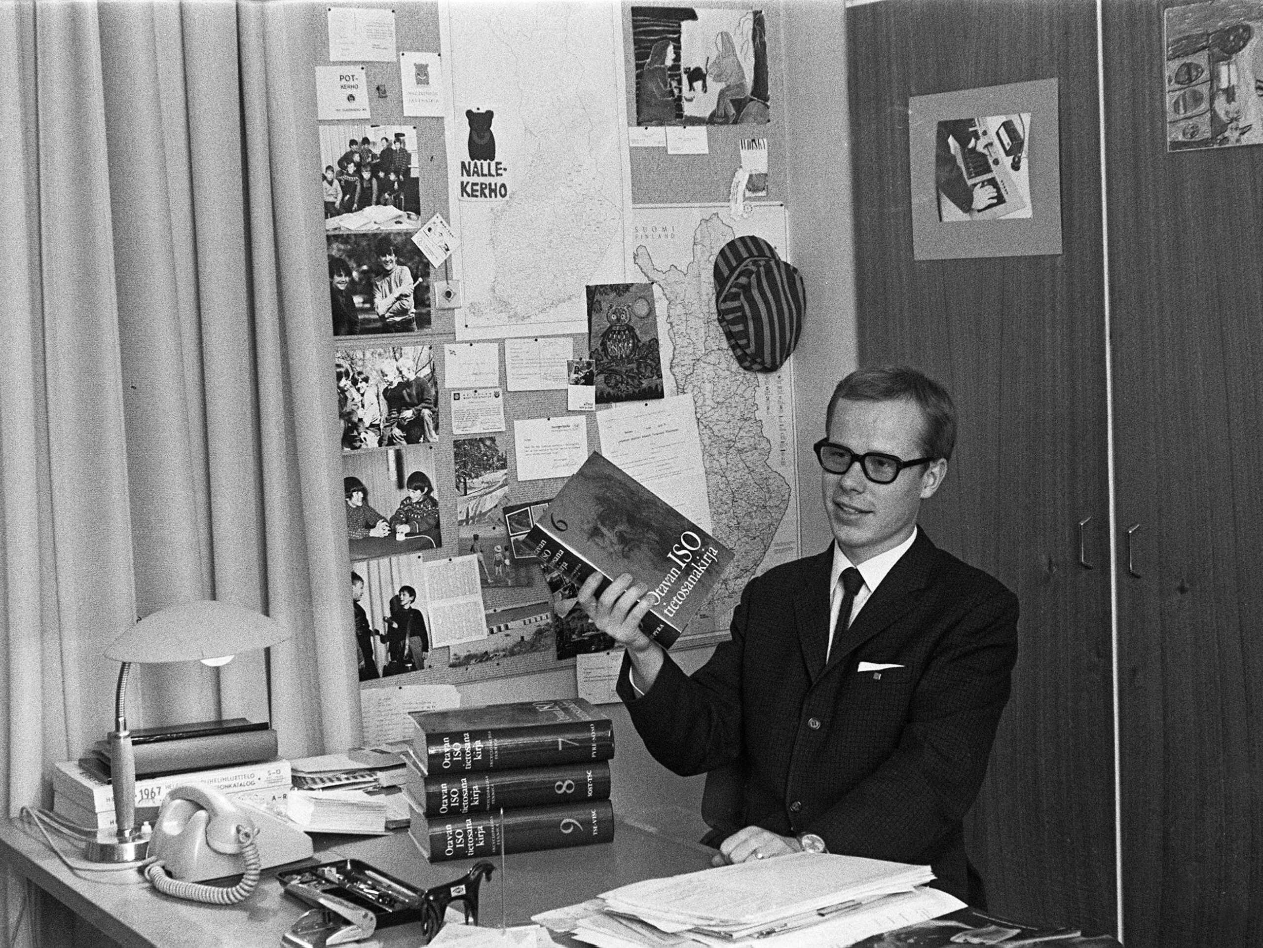 Jorma Pilke istuu työpöytänsä ääressä Yleisradion Nuorten radiossa 1960-luvulla. Pöydällä on papereita ja kirjoja, seinällä ilmoitustaululla kuvia ja papereita. Miehellä on tumma puku, valkoinen paita ja tumma solmio.