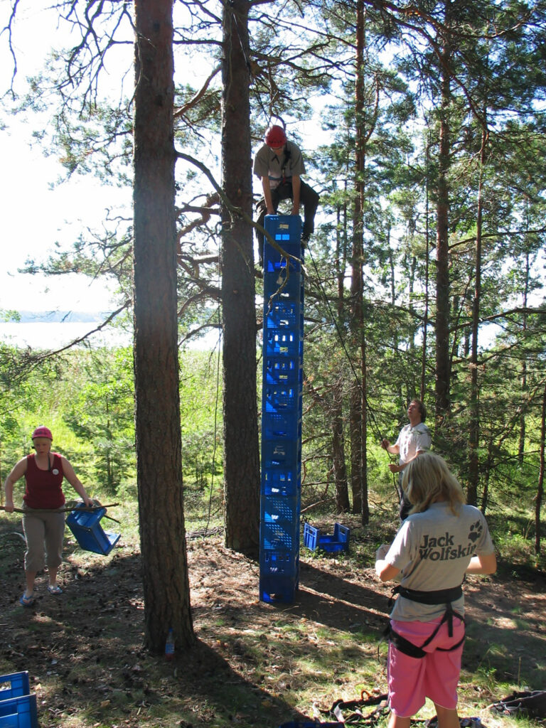 Leiriläiset rakentavat ryhmänä sinisistä muovisista (maito)laatikoista korkeaa torrnia, jonka huipulle yksi heistä on juuri kapuamassa yllään kiipeilyvaljaat ja päässään kypärä.